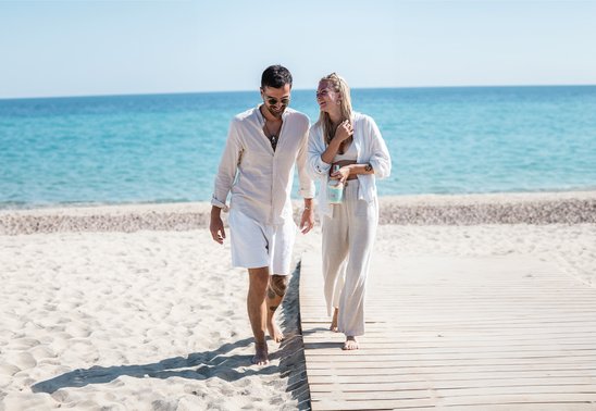 Man and woman walking along the beach laughing and holding a bottle of Liqueur Tropézienne in their hand