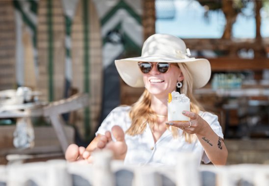 Frau mit Sonnenbrille und Hut sitzt entspannt im Beachclub und genießt Liqueur Tropézienne am Plage Pampelonne